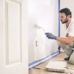 Professional painter applying white paint to interior wall, showing how long does interior paint last before you need to repaint in a residential home setting